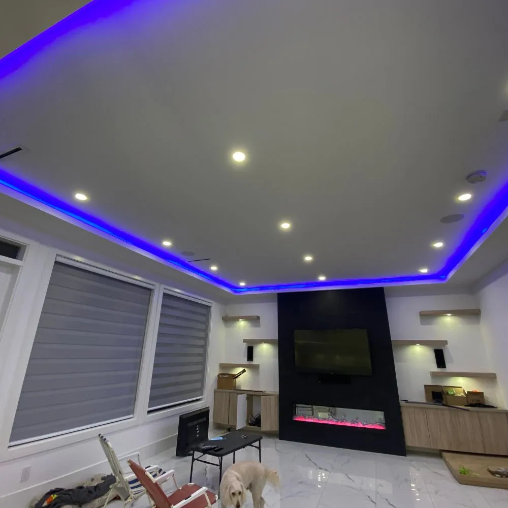 Living room with blue RGB LED strip lighting around the ceiling perimeter, recessed spotlights, a modern fireplace with a TV above it, and contemporary furniture, showcasing a clean residential RGB mood lighting installation.
