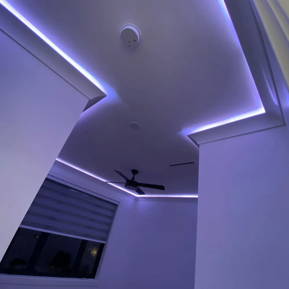 Room with soft purple RGB LED strip lighting outlining a multi angled ceiling design, creating a modern ambient glow. A ceiling fan and window with blinds are visible, highlighting a clean and minimalist RGB mood lighting setup.