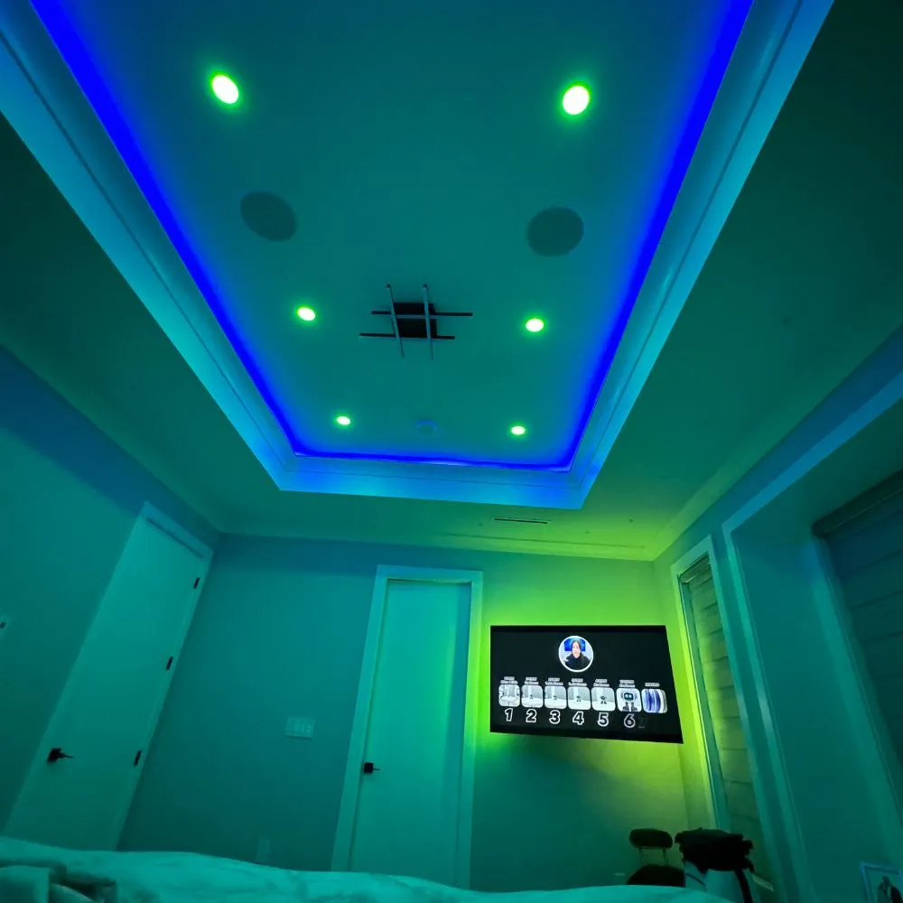 Bedroom with a tray ceiling illuminated by blue RGB LED strip lighting and green recessed lights, creating a colourful mood-lit atmosphere. A wall-mounted TV and surrounding furniture complete the modern RGB lighting setup.