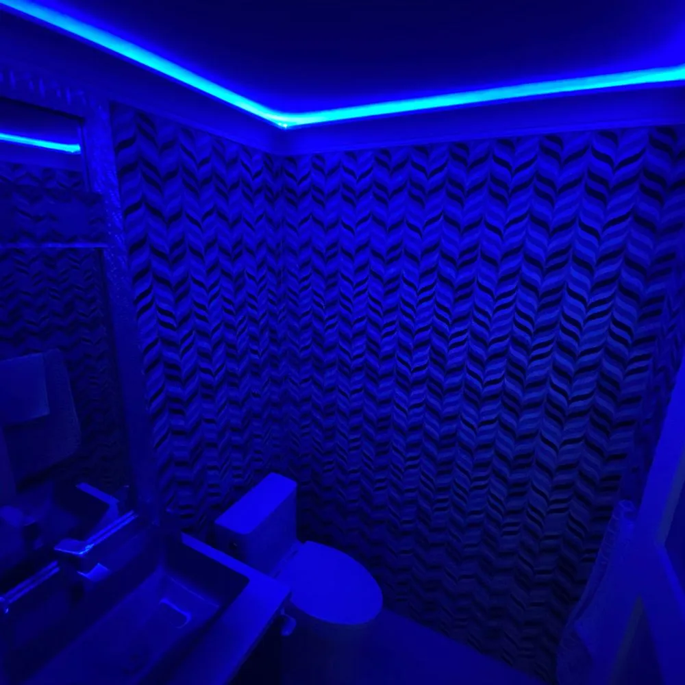 Bathroom illuminated entirely with vibrant blue RGB LED strip lighting along the ceiling perimeter, highlighting textured chevron wall tiles and modern fixtures, demonstrating a bold RGB mood lighting installation.