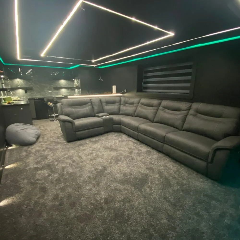 Modern media room featuring geometric LED strip lighting on the ceiling, green RGB accent lighting along the walls, a large sectional sofa, and plush carpeting, highlighting a customized RGB mood lighting installation.