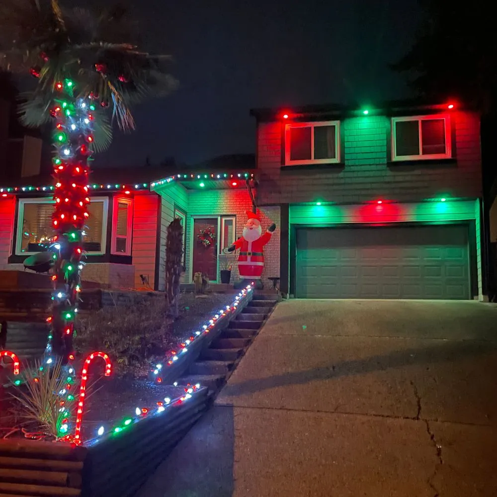 Residential home at night illuminated with vibrant RGB lighting, featuring red and green exterior accent lights along the roofline, multicolour string lights on the palm tree and pathway, and festive décor, showcasing a colourful RGB mood lighting installation.
