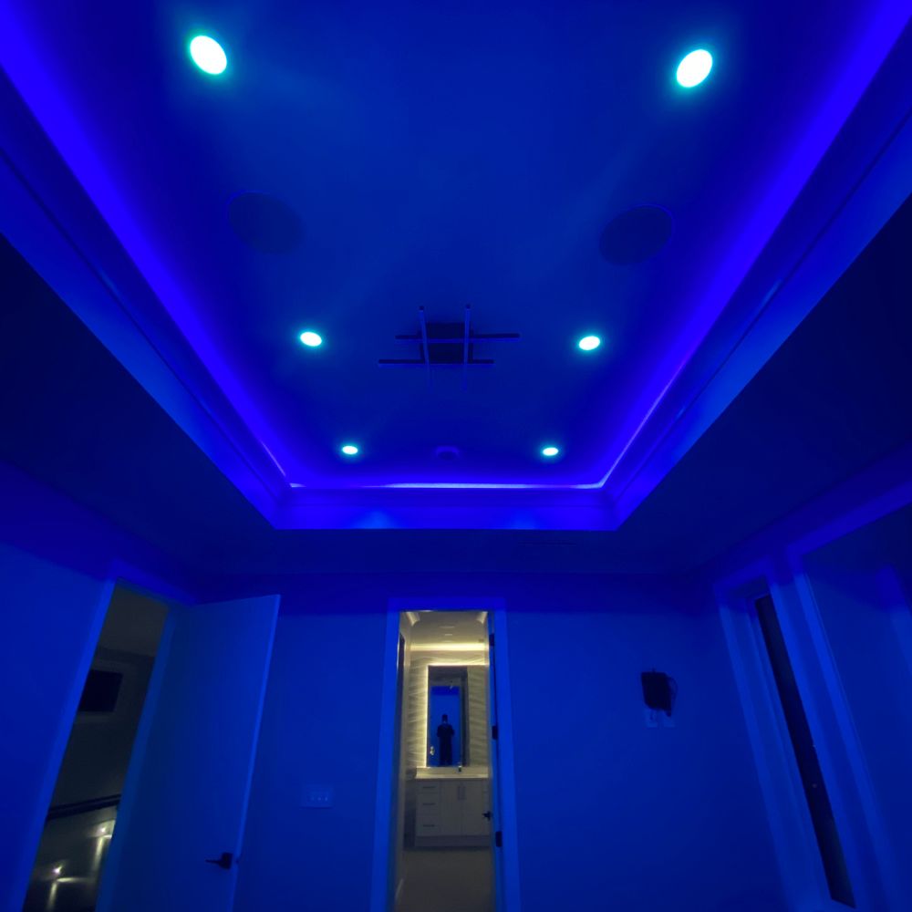 Room illuminated with deep blue RGB LED strip lighting along a tray ceiling, paired with turquoise recessed lights, creating a bold and immersive mood lighting effect. A doorway and surrounding walls are softly washed in blue light.