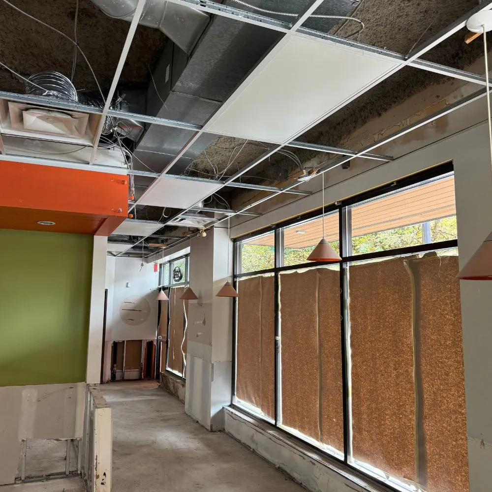 Commercial unit undergoing renovation with exposed ceiling grid, visible ductwork and electrical wiring, covered windows, and partially removed fixtures, showing the space in preparation for a commercial electrical installation upgrade.