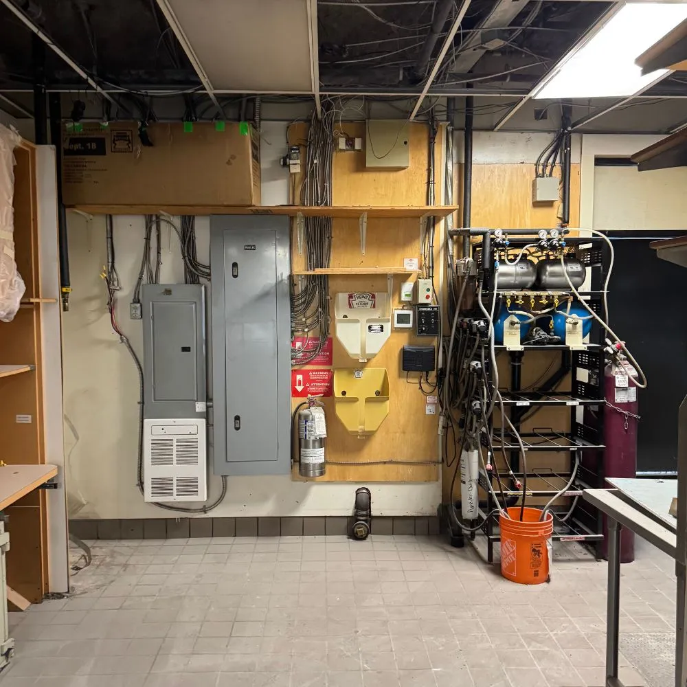 Commercial electrical room in North Vancouver with exposed wiring, mounted electrical panels, conduit runs, communication equipment, and shelving containing power and network components, showing the existing setup prior to a commercial electrical installation upgrade.