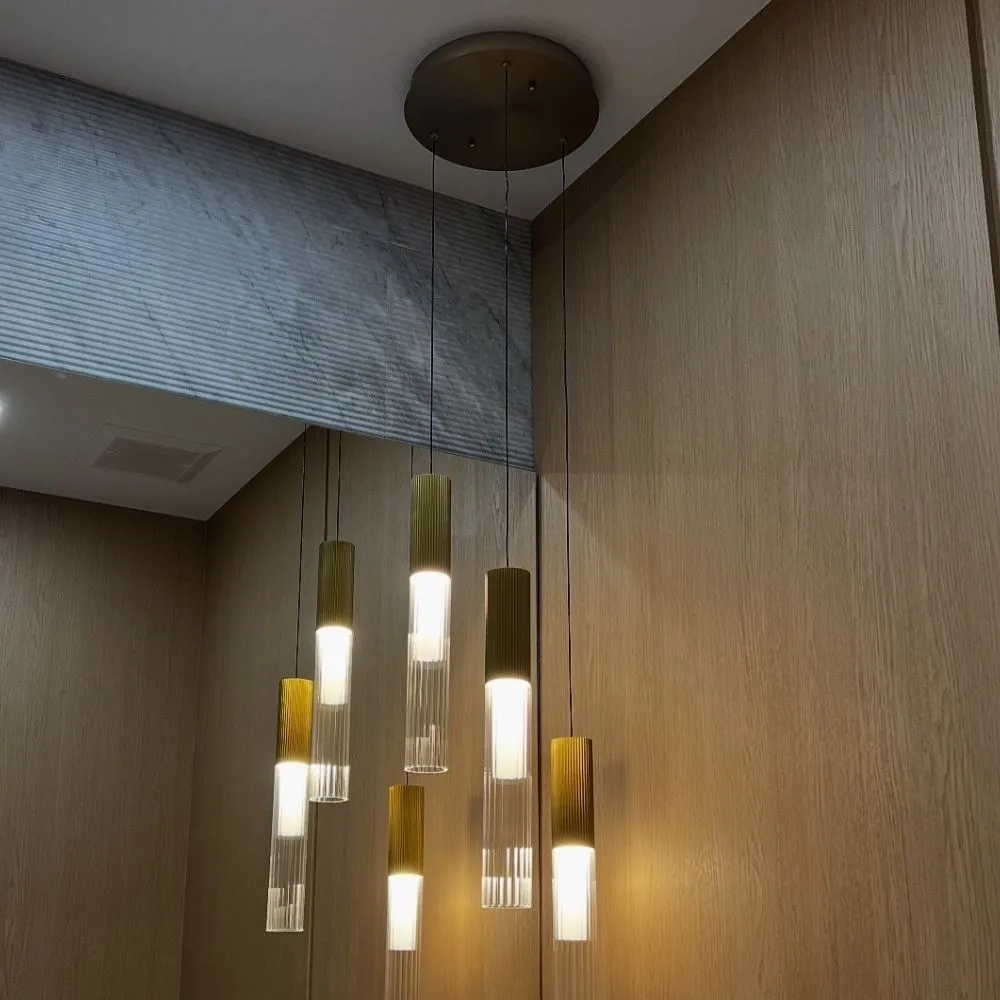 Modern multi pendant light fixture with several cylindrical gold and glass lights hanging from a round ceiling mount, installed in a corner with light wood wall panels and a grey textured ceiling feature.