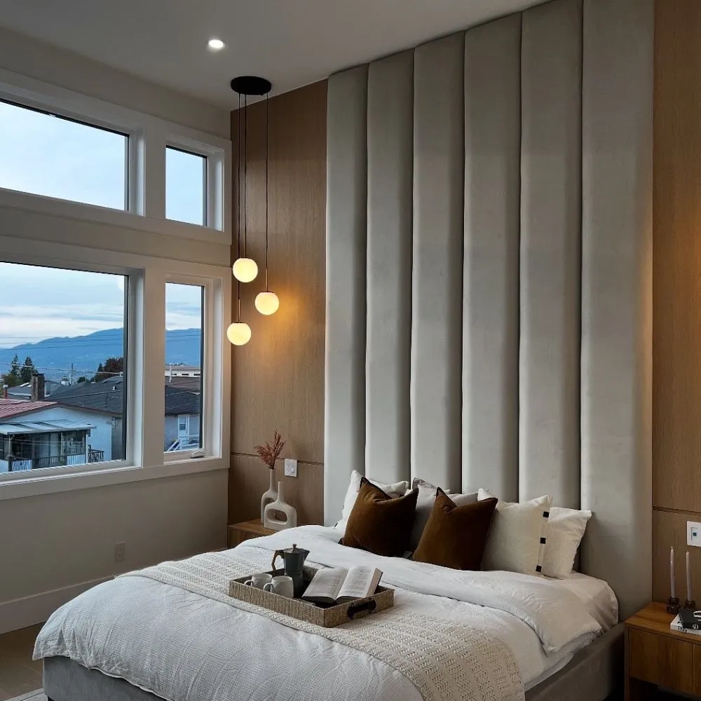 Modern bedroom with a tall upholstered headboard, three round wall-mounted globe lights on the left, large windows overlooking a residential neighborhood and mountains, and a neatly made bed with neutral linens and brown accent pillows.