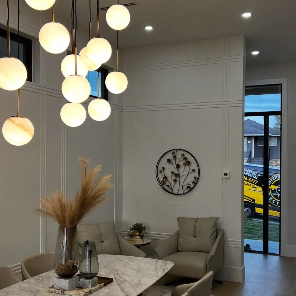 Contemporary Burnaby dining area with a cluster of hanging globe pendant lights, recessed ceiling lighting, a marble dining table, modern beige seating, and a decorative wall piece. A service van is visible through the doorway, indicating an active residential lighting installation project.