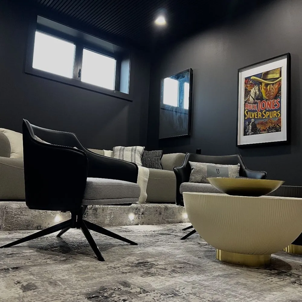 Modern home theater room with dark walls, recessed ceiling lighting, tiered seating with LED step lights, contemporary armchairs, a large sectional sofa, and framed wall art, showcasing a clean and stylish residential lighting installation.