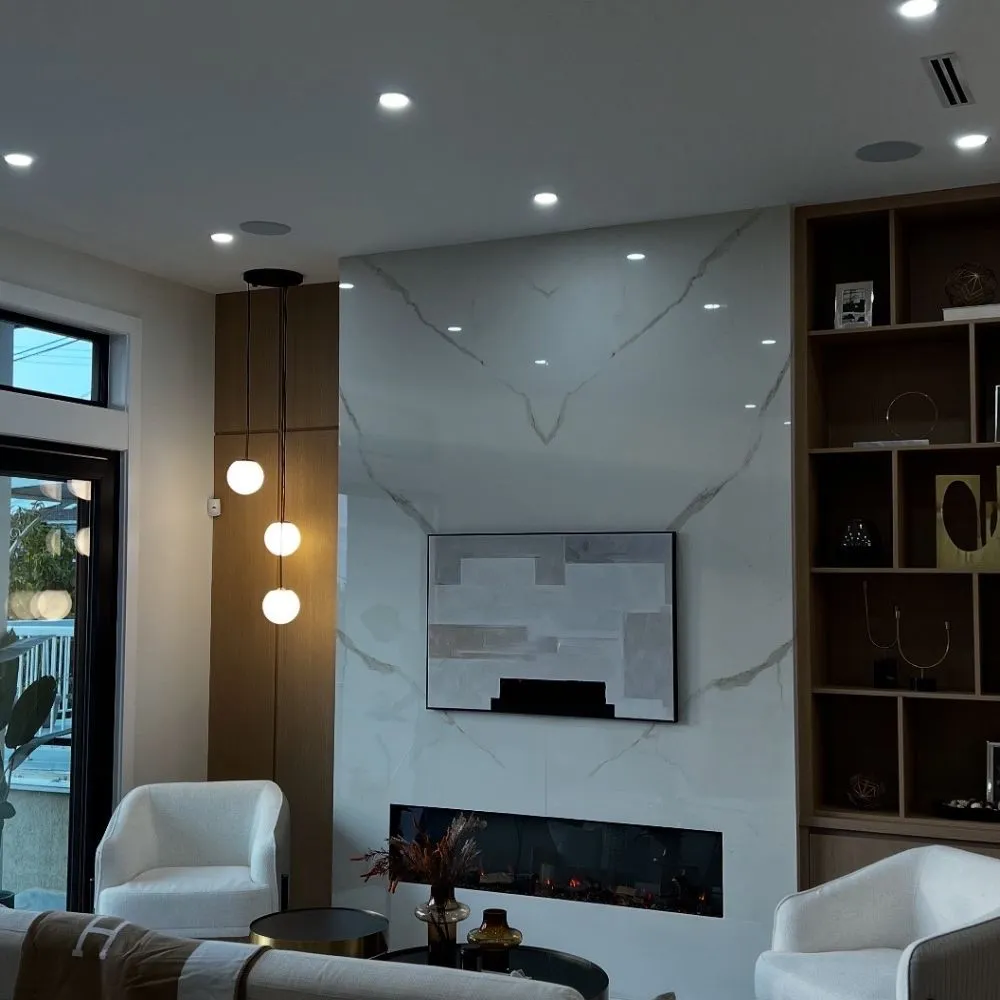 Modern living room featuring a marble accent wall with an electric fireplace, abstract artwork above it, and a trio of hanging globe pendant lights beside a tall wood panel. Two white armchairs and built-in shelving complete the contemporary space.